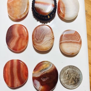 May include: A collection of polished agate stones in shades of red, orange, and white. One stone is set in a macrame pendant. A U.S. quarter is included for size reference. The stones vary in shape and pattern, with visible banding.