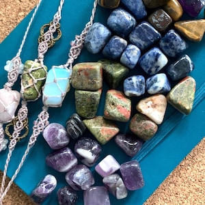 May include: An assortment of polished gemstones in various colors, including blue, green, purple, and brown, are displayed on a teal surface. Several gemstones are strung on macrame cords. The image showcases a collection of natural crystals and stones.
