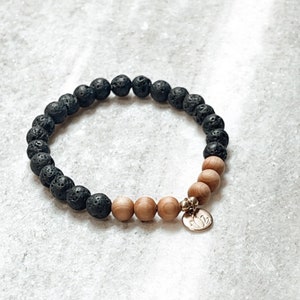 Könnte beinhalten: Ein Armband aus schwarzen Lavasteinen und Holzperlen mit einem goldenen Anhänger in Form einer Lotusblume.