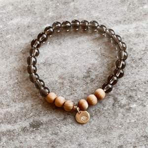 Könnte beinhalten: Ein braunes und schwarzes Perlenarmband mit einem goldenen Anhänger. Das Armband besteht aus Rauchquarz- und Sandelholzperlen.