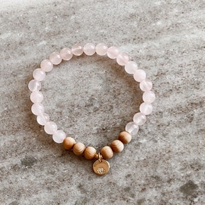Könnte beinhalten: Ein Armband aus rosa und Holzperlen mit einem goldenen Anhänger in Form einer Lotusblume.