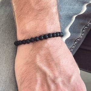 Könnte beinhalten: Ein schwarzes Lavastein-Armband. Das Armband besteht aus kleinen, runden, porösen schwarzen Perlen, die auf einem Gummiband aufgereiht sind. Das Armband wird am Handgelenk getragen.