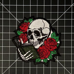 Puede incluir: Un parche bordado que muestra un cráneo humano blanco leyendo un libro verde oscuro, sostenido por una mano esquelética. El diseño está rodeado de rosas rojas vibrantes con hojas verdes, todo delineado con hilo negro sobre un fondo blanco.