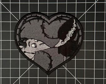 Könnte beinhalten: Ein herzförmiger Stickaufnäher mit grauem Hintergrund und schwarzen gestickten Umrissen von dornigen Ranken. Das Design zeigt zwei stilisierte Kreaturen, die sich im Profil gegenüberstehen, eine hellgrau und eine schwarz mit weißen Haar-Details.