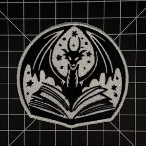 May include: A circular embroidered patch featuring a black and white design of a dragon with spread wings positioned behind an open book. The background within the circle includes a large moon and several small stars, all rendered in high contrast.