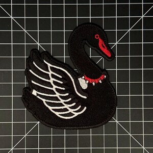 May include: An embroidered patch shaped like a black swan with white wing details, a red beak, and a red spiked collar around its neck. The swan is depicted in profile with its head curved downward, featuring bold stitching and a dark color palette.