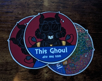 May include: Two round stickers with a black and white illustration of a ghoul wearing a gas mask and horns. The sticker has a red background with a blue banner that says "This Ghoul ate my son". The other sticker is a holographic glitter sticker.