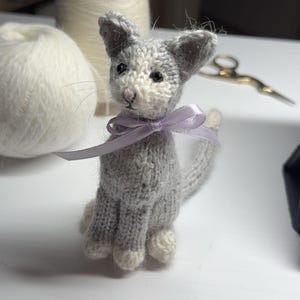 Pode incluir: Figura de gato cinzento tricotada à mão, com patas e rosto brancos. Uma fita lavanda adorna o pescoço. O gato está sentado. Lã branca e tesouras no fundo. Produto artesanal.