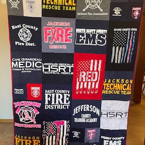 May include: A quilt made of various t-shirts featuring the logos and names of different fire and EMS departments. The quilt is made up of a grid of 16 squares, each with a different design. The quilt is black, red, white, gray, and blue.