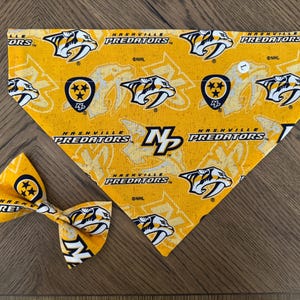 May include: A yellow and black dog bandana with the Nashville Predators logo and the text "Nashville Predators" repeated throughout. The bandana is tied around a matching bow tie.