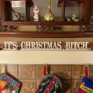 Può includere: Uno scaffale bianco con una cornice in legno e un cartello che recita "IT'S CHRISTMAS BITCH". Ci sono tre calze appese a ganci sotto lo scaffale. Le calze sono decorate con un pupazzo di neve, un design rosso e nero e Babbo Natale.
