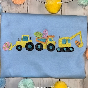 Könnte beinhalten: Ein hellblaues T-Shirt mit einem gestickten Design aus drei gelben Baumaschinen, die bunte Ostereier transportieren. Die Fahrzeuge umfassen einen Bulldozer, einen Kipplaster und einen Bagger.