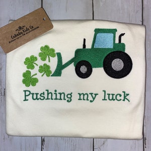 May include: Cream-colored shirt with a green tractor design pushing shamrocks. The text "Pushing my luck" is embroidered in green below the tractor. A brown tag with text is in the upper left corner.