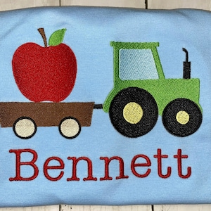 May include: A blue t-shirt featuring a green tractor pulling a wagon with a red apple inside. The name "Bennett" is embroidered in red below the tractor.