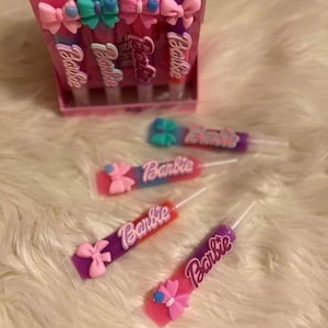 May include: A pink box set of Barbie lip gloss tubes with colourful bows. The tubes are arranged in a box and also scattered on a white, fluffy surface. Each tube has the word "Barbie" printed on it.