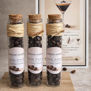 Könnte beinhalten: Drei Glasfläschchen gefüllt mit Kaffeebohnen, jeweils mit einem Korken und Bast verschlossen. Die Fläschchen tragen Etiketten mit der Aufschrift "Espresso Martini, Final Touch, A Tini Thing". Im Hintergrund ein gerahmter Druck eines Espresso Martini Cocktails.