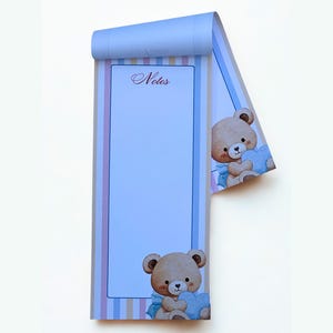 May include: A vertical notepad with a light blue background and a striped border in pastel colours. The word Notes is printed at the top in a script font. A cartoon teddy bear holding a blue heart is illustrated at the bottom and top right.