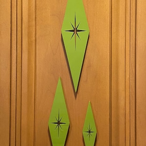 May include: Three green diamond-shaped wall decorations with a starburst cutout in the center. The decorations are different sizes and are mounted on a wood panel.