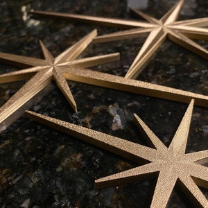 May include: Three gold-colored, star-shaped ornaments with a textured surface. The stars are arranged in a staggered pattern, with the largest star in the back and the smallest star in the front.