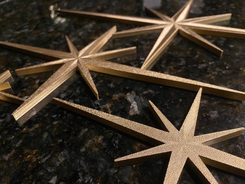 May include: Three gold-colored, star-shaped ornaments with a textured surface. The stars are arranged in a staggered pattern, with the largest star in the back and the smallest star in the front.