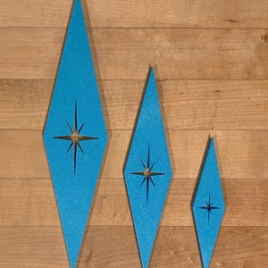May include: Three blue diamond-shaped ornaments with a starburst design in the center. The ornaments are different sizes and are arranged in a row on a wood background.