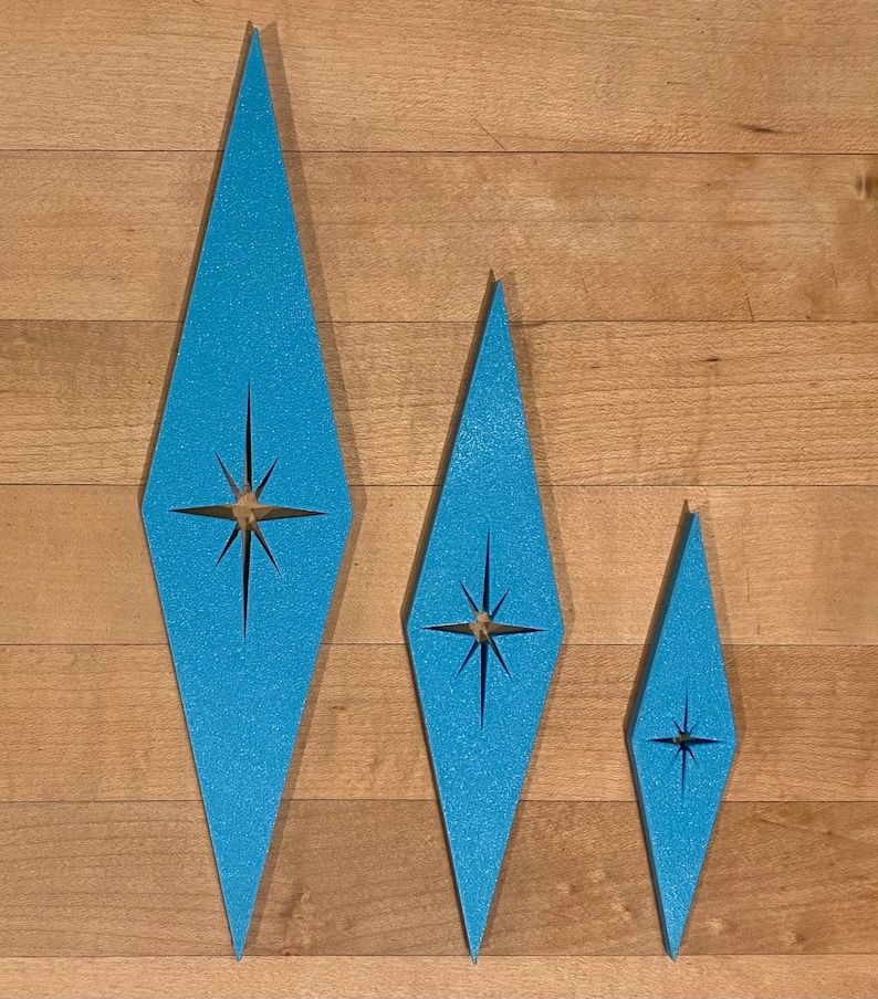 May include: Three blue diamond-shaped ornaments with a starburst design in the center. The ornaments are different sizes and are arranged in a row on a wood background.