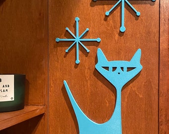 May include: A light blue cat and two light blue starburst shapes are mounted on a wood panel. The cat is stylized with a long, thin body and a long tail.