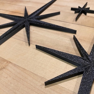 May include: Three black star-shaped decorations with a sparkly finish. The stars are made of a thick, textured material and are arranged on a light wood surface.