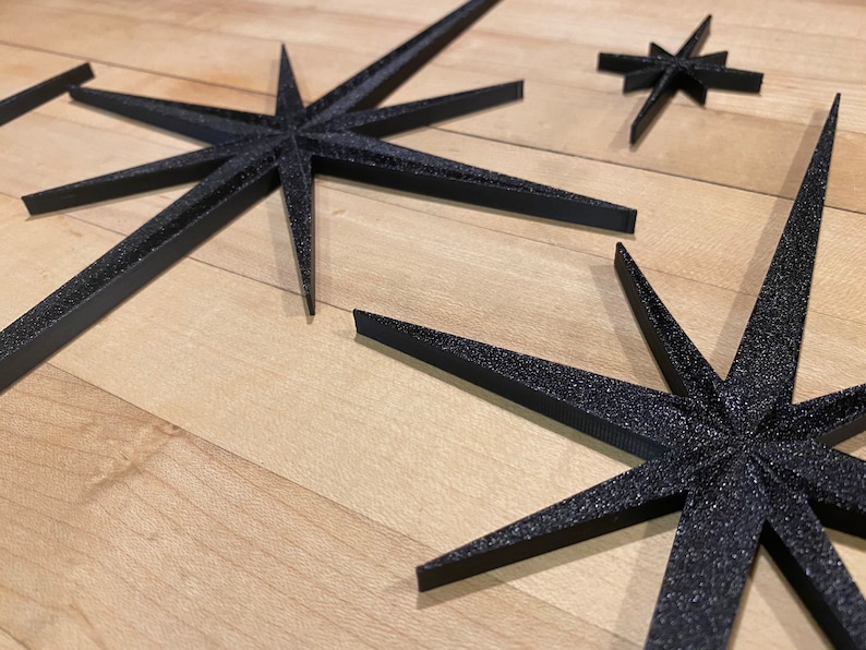 May include: Three black star-shaped decorations with a sparkly finish. The stars are made of a thick, textured material and are arranged on a light wood surface.