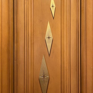 May include: Three gold diamond-shaped cabinet pulls with a starburst design in the center. The pulls are attached to a wooden cabinet door.