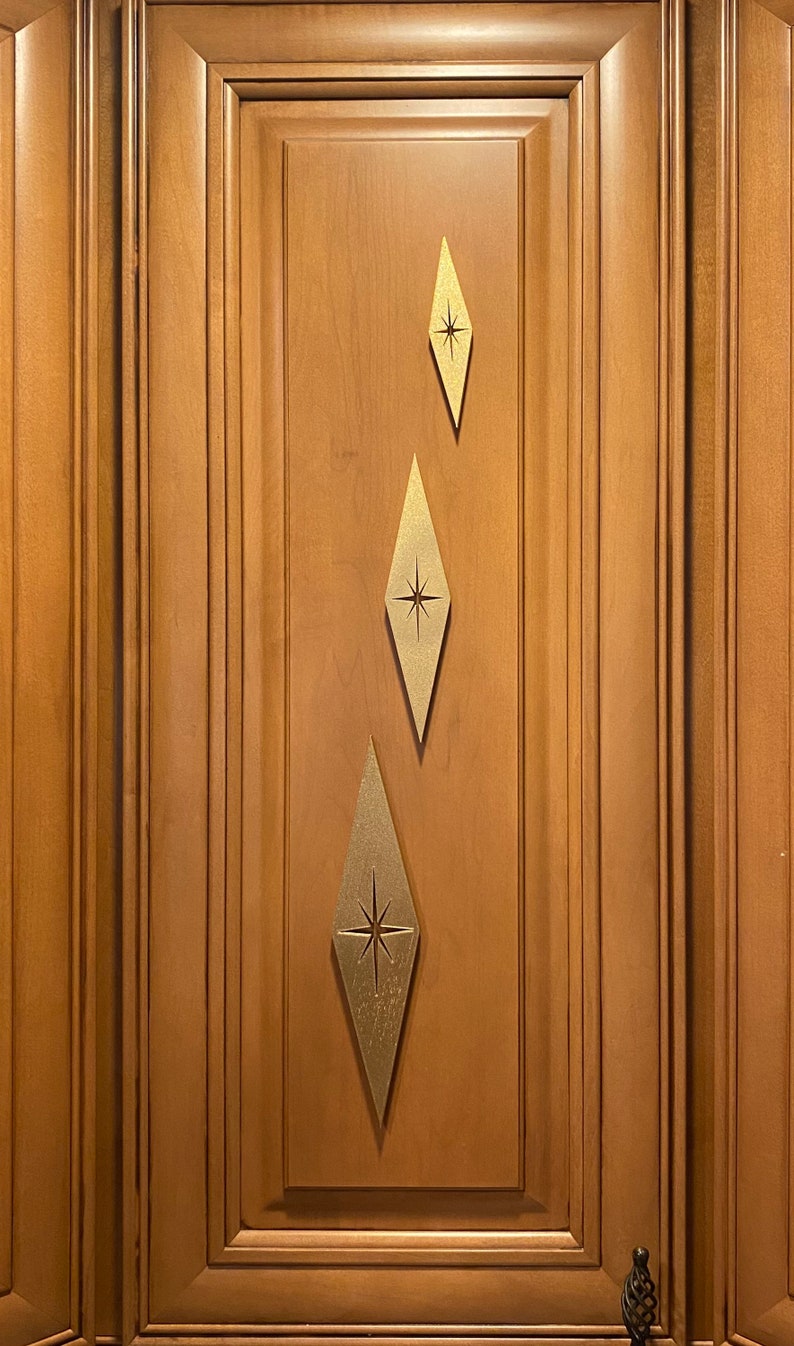 May include: Three gold diamond-shaped cabinet pulls with a starburst design in the center. The pulls are attached to a wooden cabinet door.