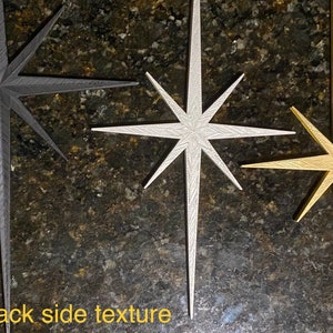 May include: Three decorative star-shaped ornaments in black, silver, and gold. The stars have a textured surface and a pointed design.
