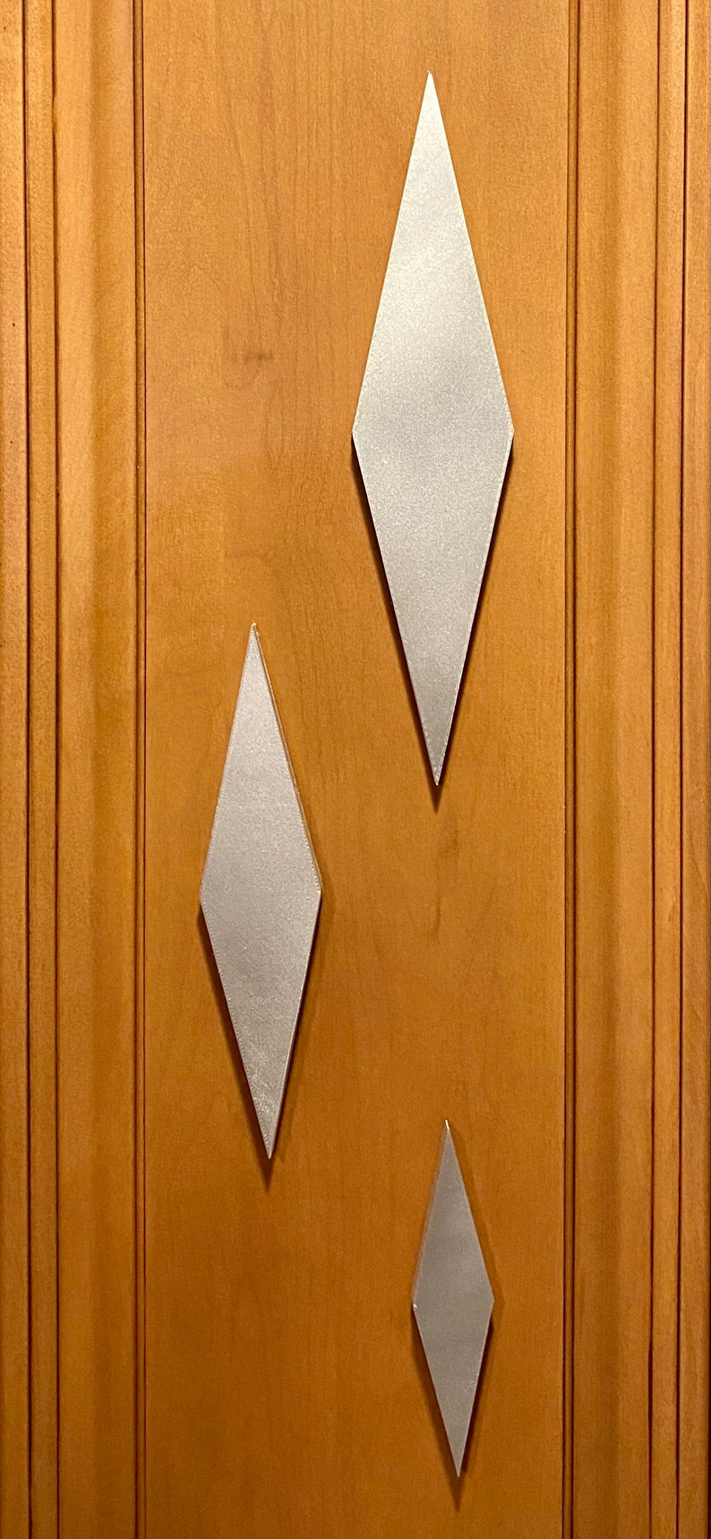 May include: Three silver diamond-shaped wall decor pieces on a wood panel background. The diamonds are different sizes, with the largest at the top and the smallest at the bottom.
