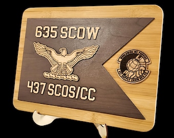 May include: A rectangular wooden plaque featuring a dark brown triangular inlay on a light bamboo base. It displays the text 635 SCOW and 437 SCOS/CC, an eagle emblem, and a circular patch with a globe graphic and the text WORLDWIDE WE PROVIDE 437 SUPPLY CHAIN OPS SQ.