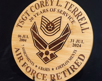 Puede incluir: Una placa redonda de madera con un acabado de madera de grano marrón. La placa presenta un diseño grabado con láser de un emblema de la Fuerza Aérea, el texto "TSGT COREY L. TERRELL", "20 YEARS OF SERVICE", "06 JUL 2004", "31 JUL 2024", "7 ASSIGNMENTS", "6 BASES", "4 DEPLOYMENTS" y "AIR FORCE RETIRED".