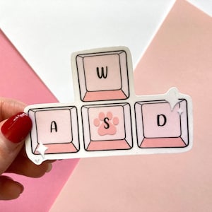 Puede incluir: Pegatinas de teclas de teclado rosa y blancas con las letras W, A, S y D. La tecla S tiene una huella de pata en lugar de una letra.