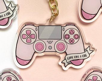 May include: Pink acrylic keychain featuring a video game controller design. The controller has pink buttons and a gray center, with the text "GAME LIKE A GIRL" on a banner. A gold chain is attached for hanging.