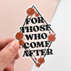 Op de afbeelding: Een witte sticker met de tekst "FOR THOSE WHO COME AFTER" in een zwarte ruit, versierd met oranje rozen en groene wijnranken. De sticker wordt door een hand vastgehouden tegen een roze en witte achtergrond.