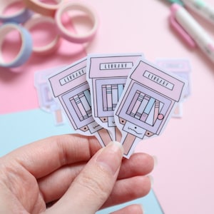 Peut inclure: Trois autocollants roses et blancs avec le mot "LIBRARY" imprimé dessus. Chaque autocollant présente une image de bibliothèque en dessin animé avec des livres colorés.