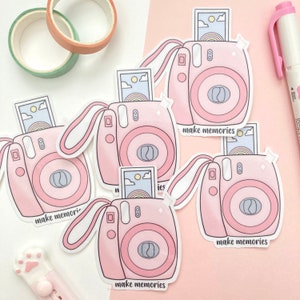 May include: Four pink and white instant camera stickers with a photo inside each camera. Each sticker has the text "make memories" printed on it.