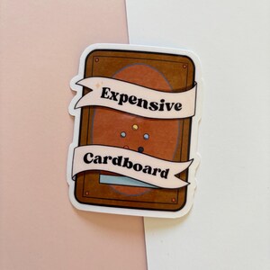 Puede incluir: Una pegatina con fondo marr&oacute;n y una pancarta blanca que dice "Expensive" y "Cardboard". La pegatina tiene un borde blanco y est&aacute; sobre un fondo rosa y blanco.