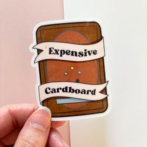 Puede incluir: Una pegatina con un dise&ntilde;o marr&oacute;n y beige, con el texto "Expensive Cardboard" en una pancarta blanca. La pegatina tiene un acabado brillante y est&aacute; sujeta por una mano sobre un fondo rosa y blanco.