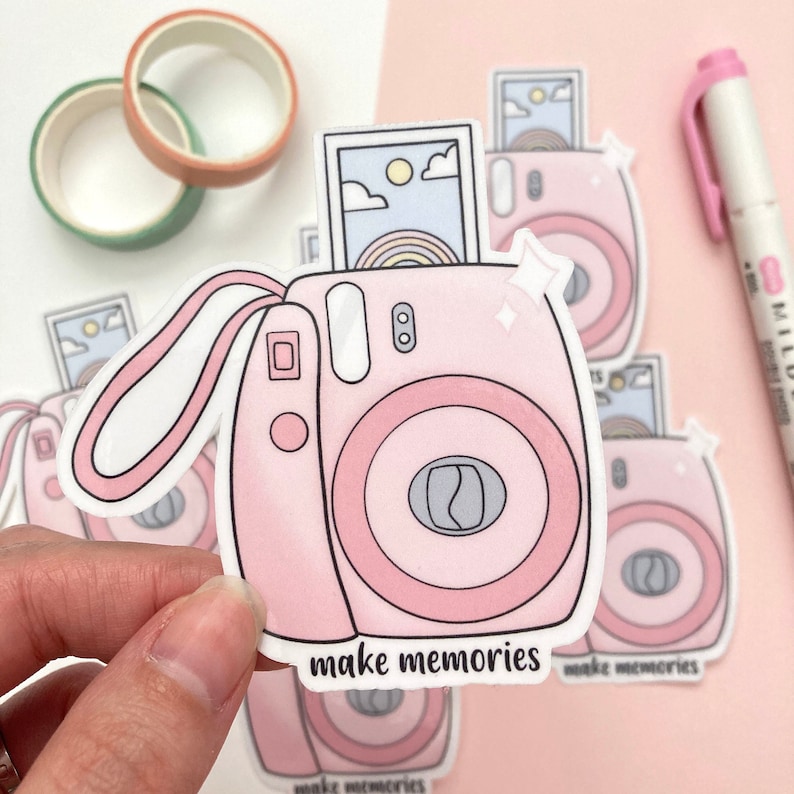 May include: Pink and white sticker of a cartoon instant camera with a rainbow photo inside. The sticker says "make memories".