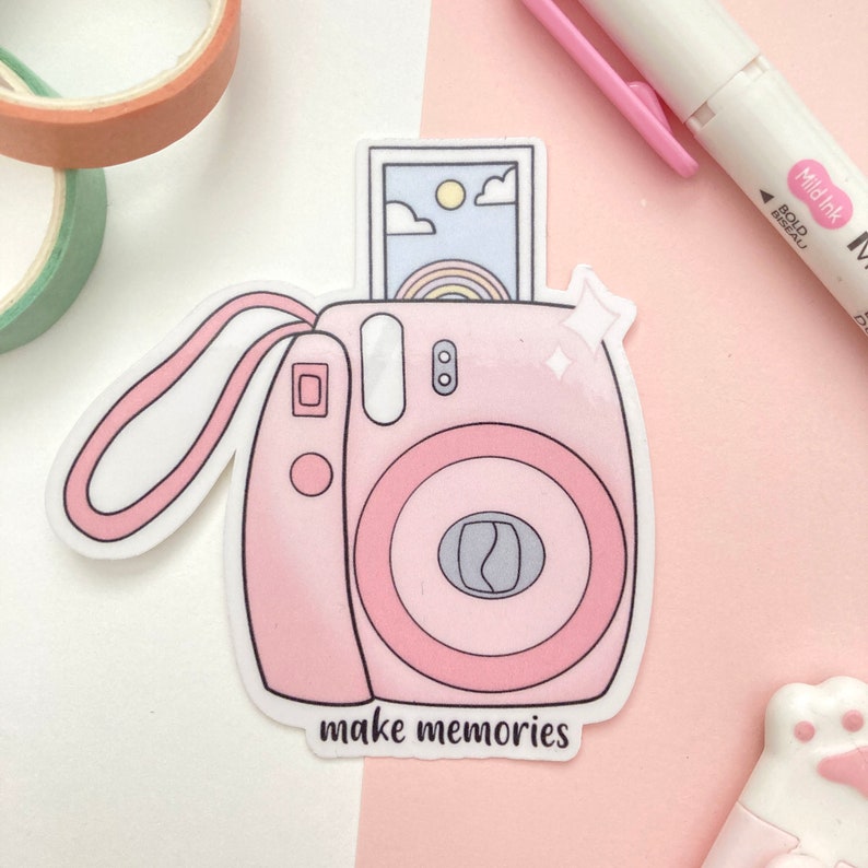 May include: Pink and white cartoon illustration of an instant camera with a photo inside. The camera has a strap and the text "make memories" is below the camera.