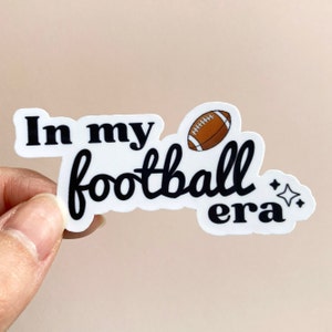 Puede incluir: Pegatina blanca con texto negro que dice "In my football era" con un gráfico de fútbol marrón y dos pequeñas estrellas blancas.