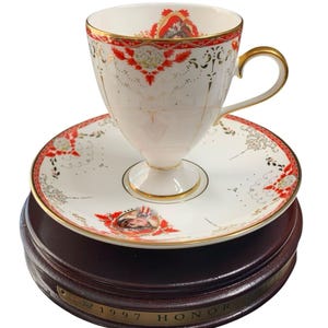 May include: A white teacup and saucer set with red and gold floral designs. The teacup has a gold handle and rim. The saucer and teacup sit on a wooden base with a gold plaque that reads "1997 HONOR".