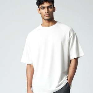May include: A white crew neck t-shirt with short sleeves. The shirt has a loose fit and is made of a textured material. The model is wearing dark grey trousers. The overall style is casual and contemporary.