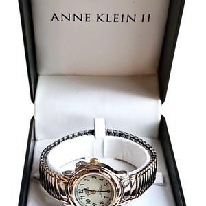 May include: Anne Klein II watch in a black box. The watch has a silver and gold-tone expanding band and a white face with black numerals. The watch is displayed in a black box with the brand name printed in white.
