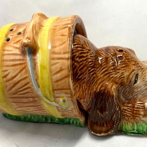 Può includere: Un set di saliera e pepiera in ceramica raffigurante un cane marrone che esce da una botte di legno. La botte ha fasce gialle e un manico argentato. Il cane poggia su una base verde, con fori per sale e pepe.