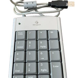 Puede incluir: Un teclado numérico Targus plateado con teclas grises y un cable USB. El teclado tiene un diseño estándar con teclas numéricas, teclas de función y un indicador de Bloq Num. El logotipo de Targus es visible en la parte superior.